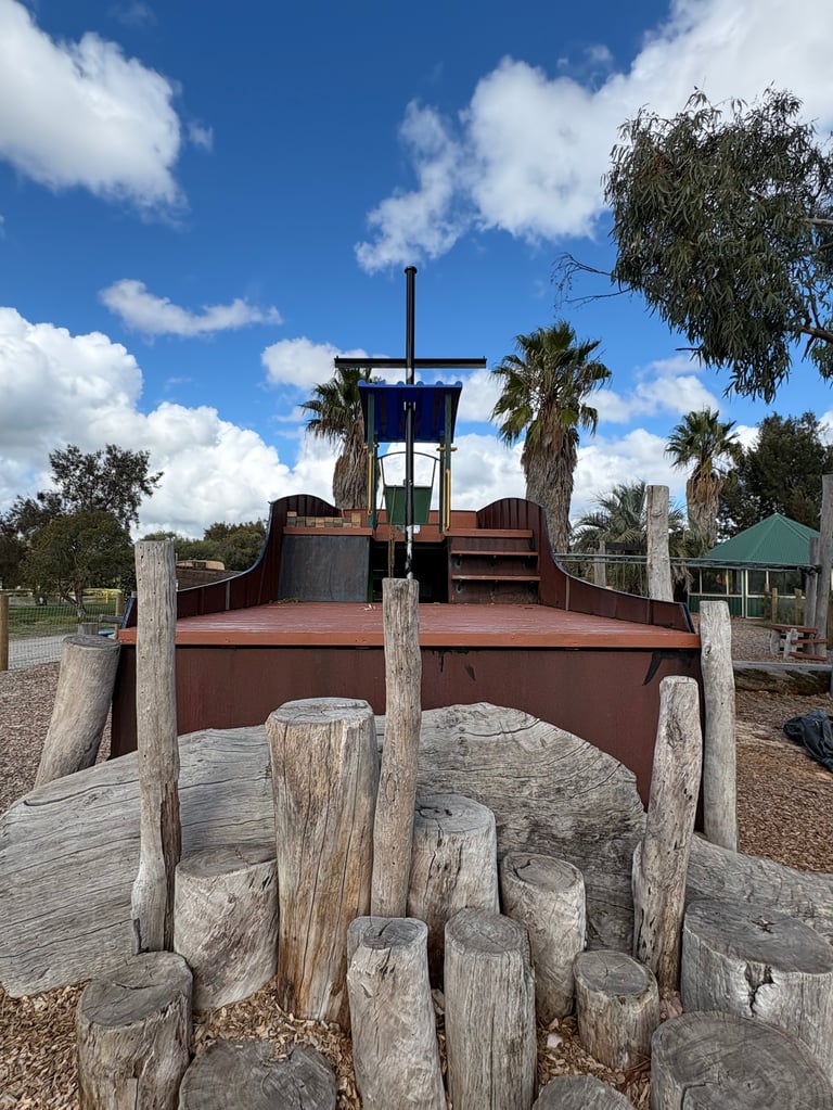 Wooden playground shaped like a pirate ship with log stumps in foreground, palm trees and blue sky background
