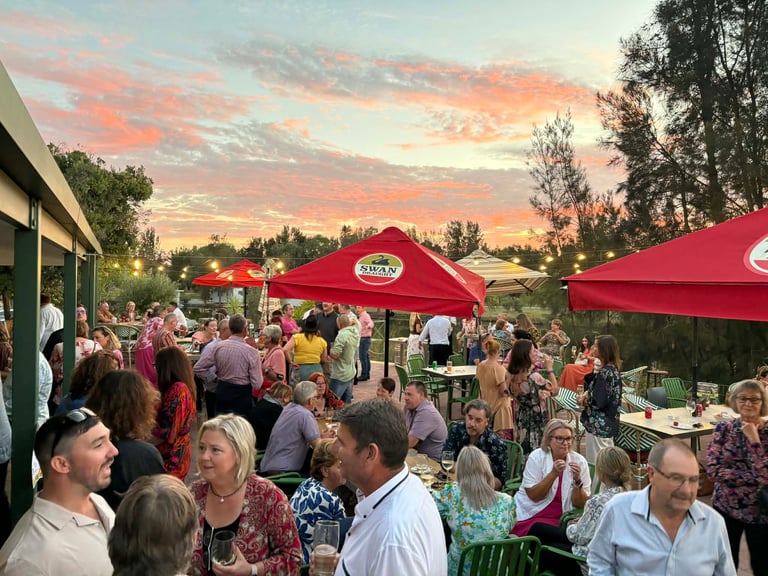 Outdoor crowd gathered under red umbrellas at sunset event with buildings and trees in background