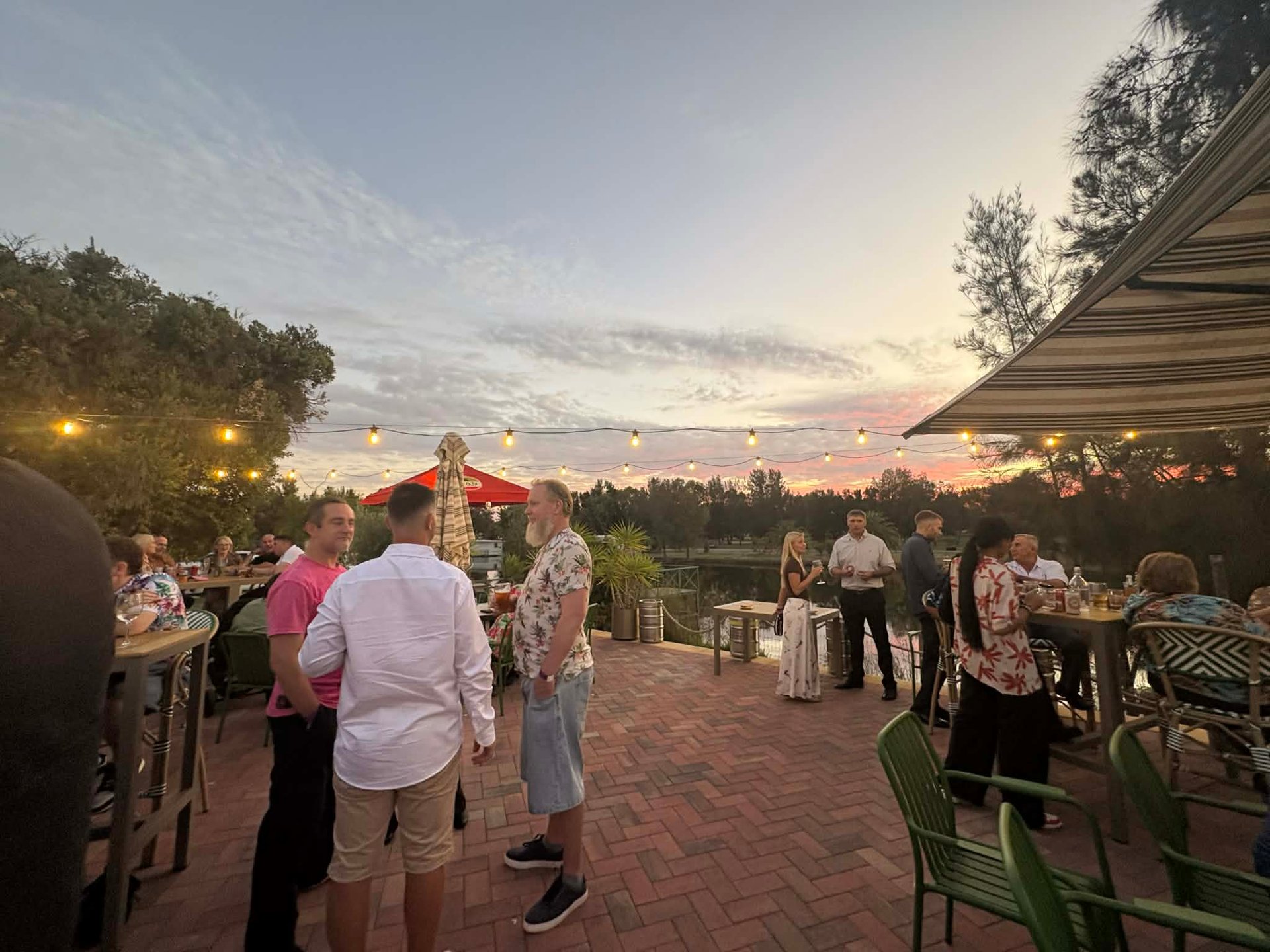 Outdoor evening event with guests mingling on a brick patio at sunset under an awning with trees in the background