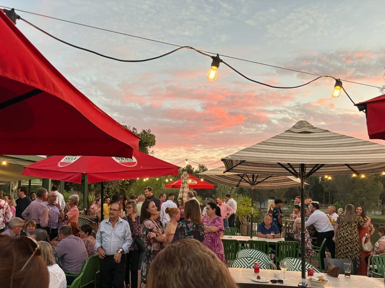 Outdoor garden party at sunset with red and white umbrellas, string lights, and guests mingling under a colorful pink and orange sky
