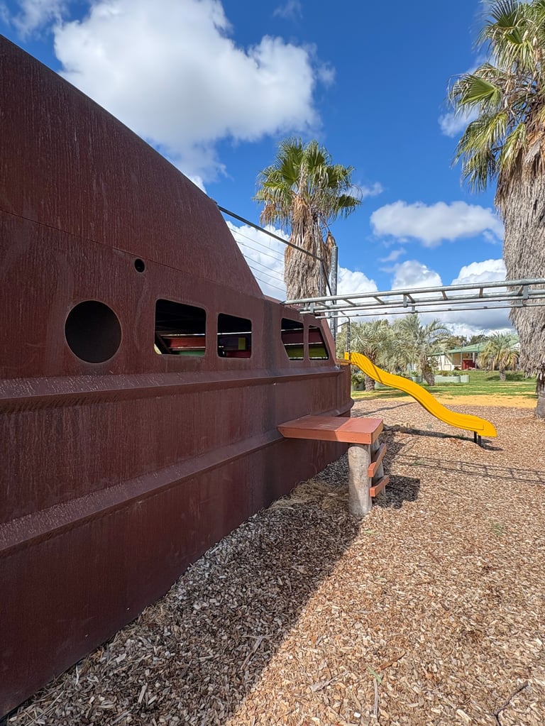 Rusty red metal playground equipment shaped like a boat with round windows and yellow slide, surrounded by palm trees and gravel