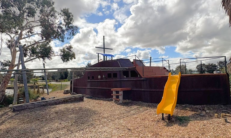 Rusty boat-shaped playground with yellow slide, cross on top, in sandy yard with trees and blue sky