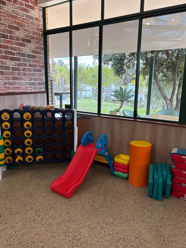 Indoor play area with colorful climbing equipment, red slide, stacked foam blocks, and large windows overlooking park grounds